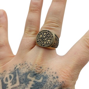 A gold-toned ring with a circular design featuring a star with eight points and the letters "Azazel" around the perimeter. The ring also has a triangular design on the band.
