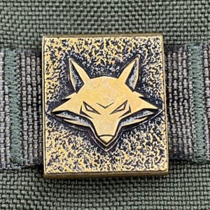 Peut inclure: Boucle carrée en métal couleur laiton avec un motif détaillé de tête de renard. La tête de renard est centrée sur la boucle, avec un fond texturé. La boucle est fixée à un tissu rayé vert et gris.