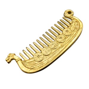 A gold-colored metal comb shaped like a Viking longship Drakkar. The comb features a detailed design of a ship with circular embellishments along the side and a dragon head at the front. The comb's teeth are evenly spaced.