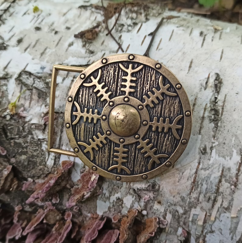 May include: A gold-toned belt buckle with a circular design featuring a shield with a central circle and a pattern of stylised branches. The buckle is on a birch bark background.