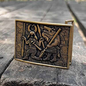 May include: A rectangular brass-toned metal belt buckle featuring a relief design of a horned figure holding a spear and a wolf-like creature holding a staff. The surface has a textured, weathered appearance with dark recessed details and raised edges.
