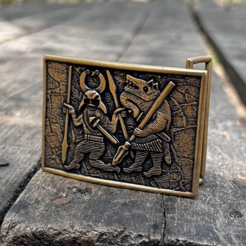 May include: A rectangular brass-toned metal belt buckle featuring a relief design of a horned figure holding a spear and a wolf-like creature holding a staff. The surface has a textured, weathered appearance with dark recessed details and raised edges.