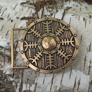 May include: A gold-toned metal belt buckle with a circular design featuring a stylised shield with a central circle and radiating lines. The shield is surrounded by a decorative border.