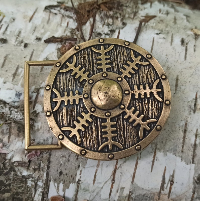 May include: A gold-toned metal belt buckle with a circular design featuring a stylised shield with a central circle and radiating lines. The shield is surrounded by a decorative border.