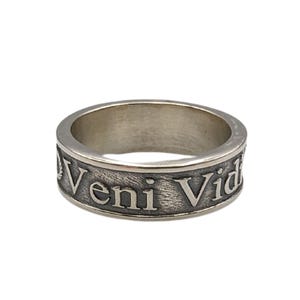 May include: A silver-toned metal ring with a textured, darkened background. The band features the embossed Latin text Veni Vici, with a small laurel wreath symbol visible at the start of the inscription. The ring has a smooth, polished interior.