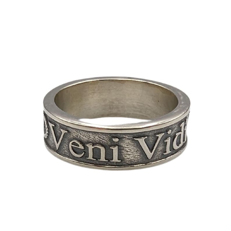 May include: A silver-toned metal ring with a textured, darkened background. The band features the embossed Latin text Veni Vici, with a small laurel wreath symbol visible at the start of the inscription. The ring has a smooth, polished interior.