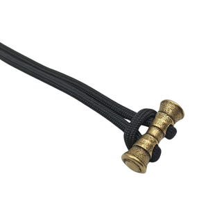 May include: A gold-coloured metal paracord toggle with a textured surface. The toggle is attached to a black paracord.