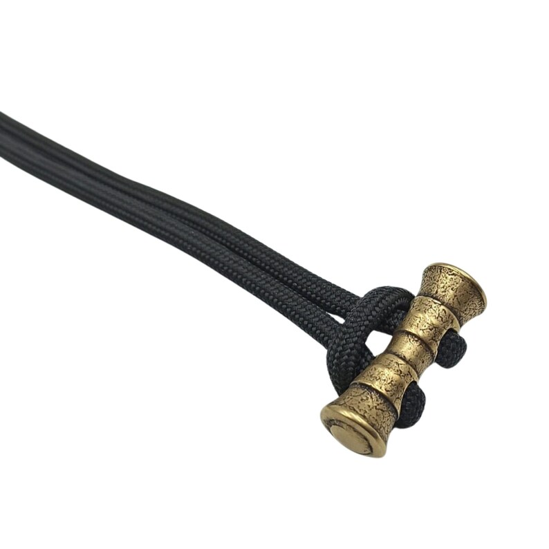 May include: A gold-coloured metal paracord toggle with a textured surface. The toggle is attached to a black paracord.