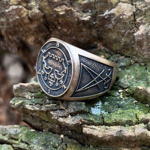 Pode incluir: Um anel de cor bronze com um design detalhado. O anel apresenta um emblema circular com símbolos intrincados e a letra "B". A banda tem um design triangular com um símbolo em forma de estrela. O anel está sobre um fundo natural e musgoso.