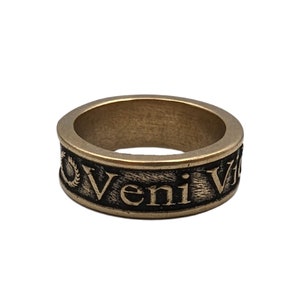 May include: A bronze-toned metal ring featuring a textured, darkened band with the embossed Latin inscription Veni Vidi Vici. The ring has a wide, flat profile with a weathered finish, showcasing raised lettering and a laurel wreath symbol.