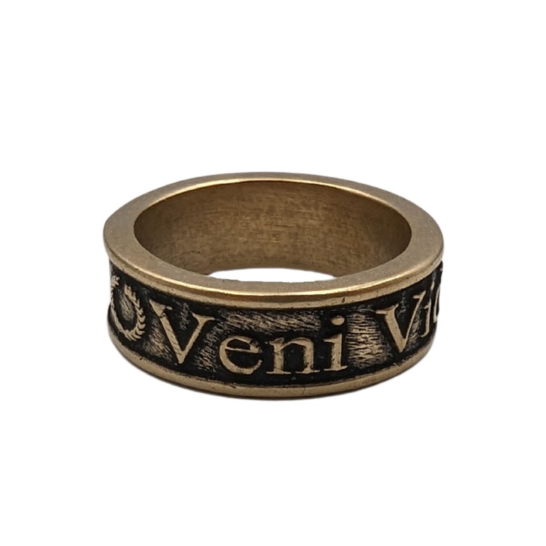 May include: A bronze-toned metal ring featuring a textured, darkened band with the embossed Latin inscription Veni Vidi Vici. The ring has a wide, flat profile with a weathered finish, showcasing raised lettering and a laurel wreath symbol.