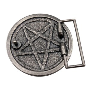 May include: A circular silver-toned metal belt buckle featuring a raised pentagram design with a textured, weathered finish. The buckle includes a rectangular frame attachment on the right side and a small metal stud on the left side for fastening.