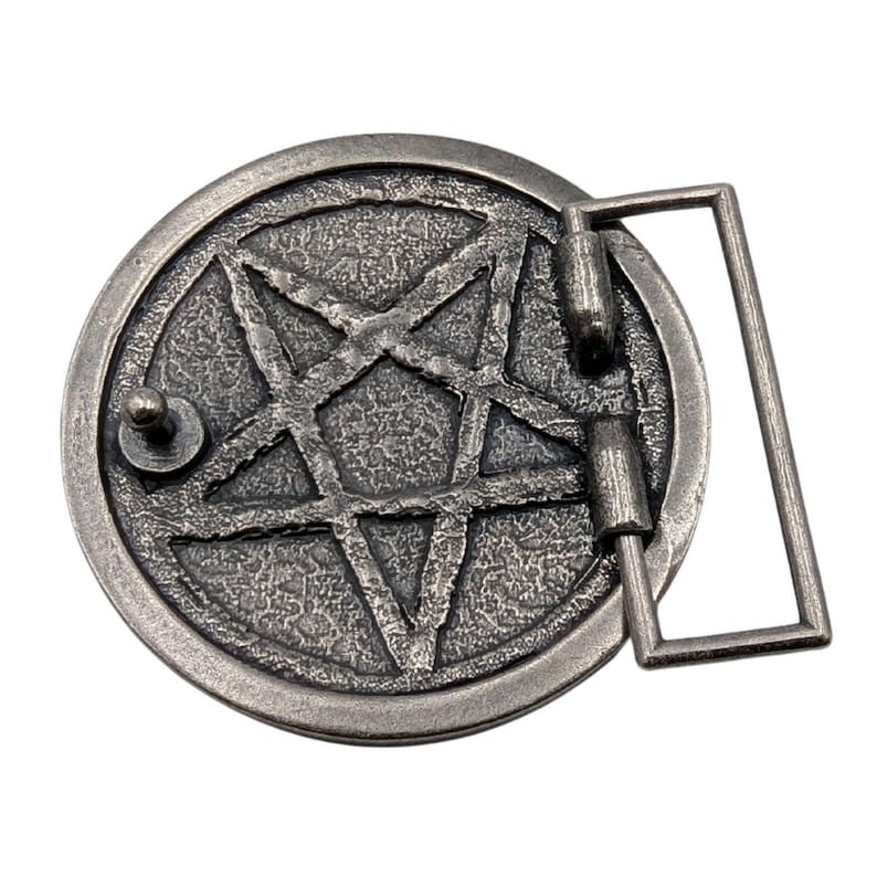 May include: A circular silver-toned metal belt buckle featuring a raised pentagram design with a textured, weathered finish. The buckle includes a rectangular frame attachment on the right side and a small metal stud on the left side for fastening.
