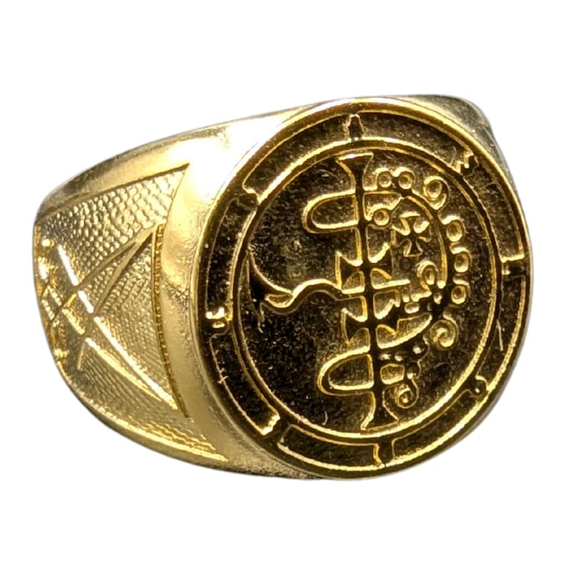 May include: A gold ring with a detailed design featuring a circular symbol with intricate lines and curves. The ring has a textured band.