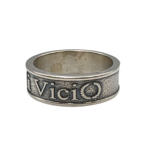 May include: A silver-toned metal ring with a textured, oxidised finish. The band features the embossed text "Vici" followed by a circular laurel wreath emblem. The interior of the band is smooth and polished, contrasting with the dark, rugged exterior surface.