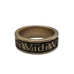May include: A metallic band ring with a brushed bronze finish featuring the engraved Latin phrase Veni Vidi Vici in black lettering. The ring has a wide, flat profile with a textured surface around the circumference where the text is displayed.