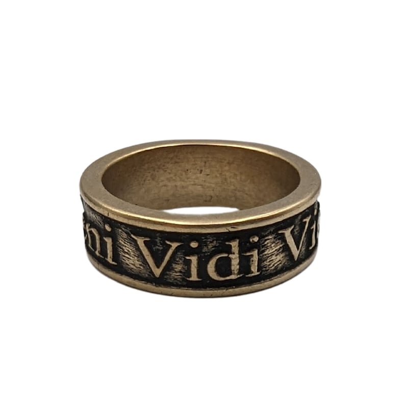 May include: A metallic band ring with a brushed bronze finish featuring the engraved Latin phrase Veni Vidi Vici in black lettering. The ring has a wide, flat profile with a textured surface around the circumference where the text is displayed.
