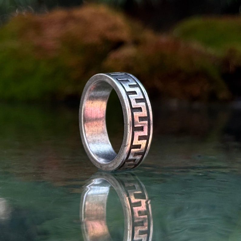 May include: A silver-toned metal ring featuring a continuous black recessed Greek key pattern engraved around the outer band. The ring has a smooth, polished interior surface and a flat profile, showcasing a distinct geometric design.