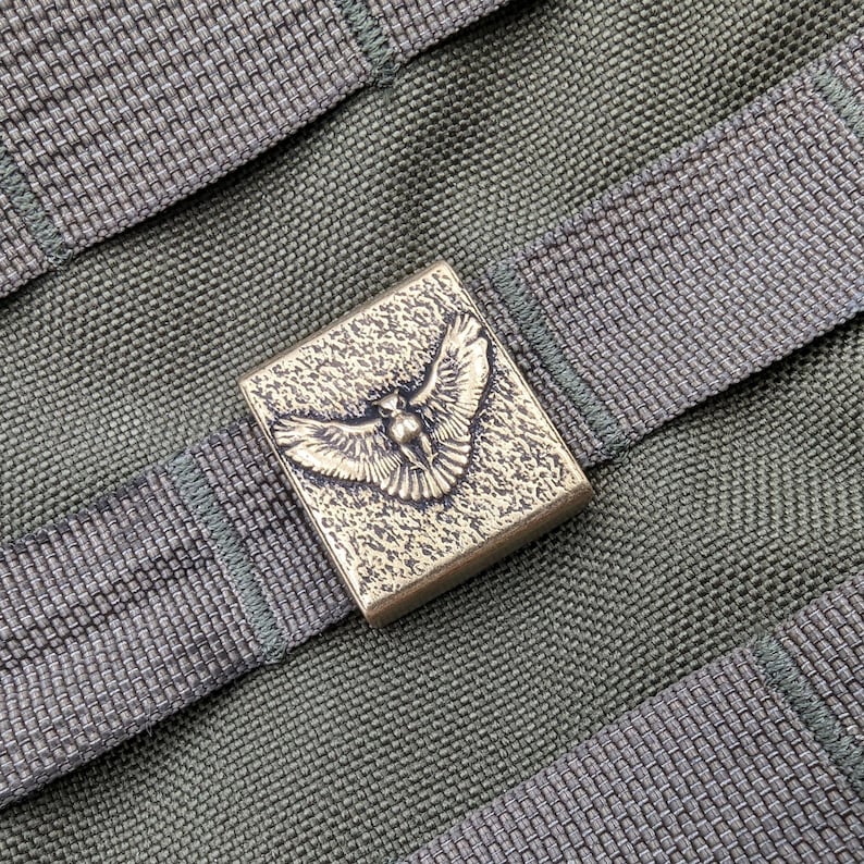 Pu&ograve; includere: Una fibbia in metallo color ottone con un design dettagliato di un uccello in volo. La fibbia &egrave; attaccata a un cinturino in tessuto verde.