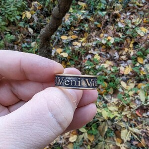 May include: A metallic band ring featuring a dark, recessed background with the raised, polished text Veni Vidi Vici engraved along the outer surface. The ring has a flat profile and a distinct contrast between the dark lettering and the metal.