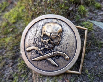 May include: A gold-toned belt buckle with a skull and crossbones design. The skull is detailed with a toothy grin and crossed bones.