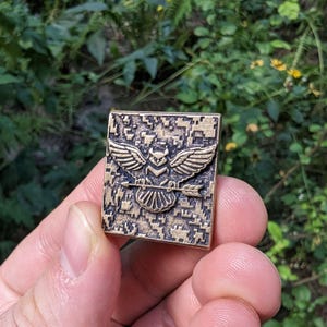 Pu&ograve; includere: Uno spillo in metallo color bronzo con un motivo mimetico pixelato. Lo spillo presenta un gufo stilizzato con le ali spiegate che tiene una freccia negli artigli.