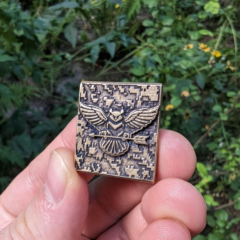 Pu&ograve; includere: Uno spillo in metallo color bronzo con un motivo mimetico pixelato. Lo spillo presenta un gufo stilizzato con le ali spiegate che tiene una freccia negli artigli.