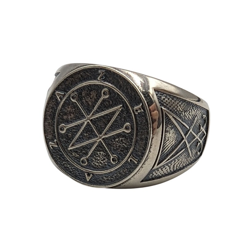 A silver ring with a circular design featuring a symbol and the letters "Azazel " around the edge. The ring has a textured band with a smaller symbol on the side.