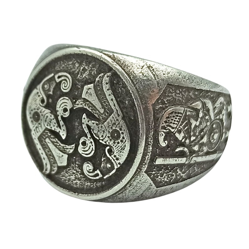 May include: A silver ring with an intricate design of two intertwined animals. The ring is oval shaped and has a textured surface.