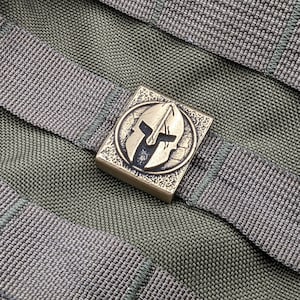 May include: A bronze-colored metal buckle with a Spartan helmet design. The buckle is attached to a green nylon strap.