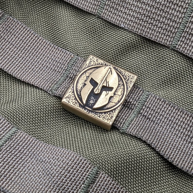 May include: A bronze-colored metal buckle with a Spartan helmet design. The buckle is attached to a green nylon strap.