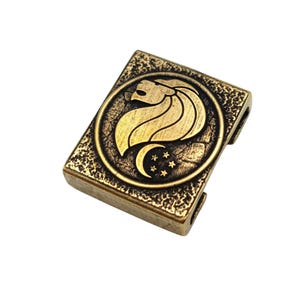 May include: A rectangular antique brass-toned metal bead featuring a textured surface and a central circular emblem. The emblem displays a stylized lion head profile above a crescent moon and five small stars, with side slots for threading.