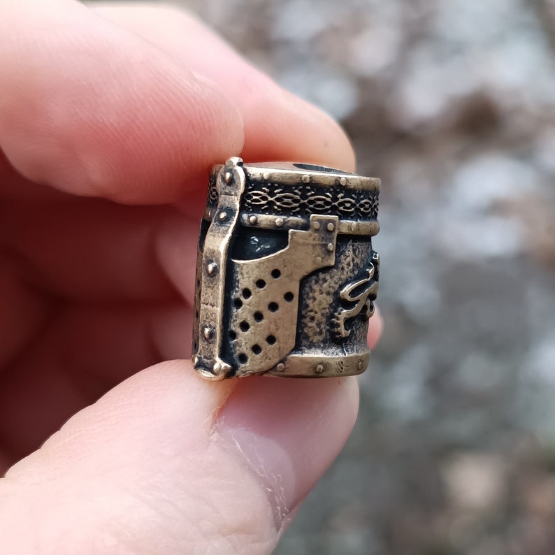May include: A gold-colored metal bead with a detailed design of a knight's helmet. The bead has a textured surface and a decorative band around the top.