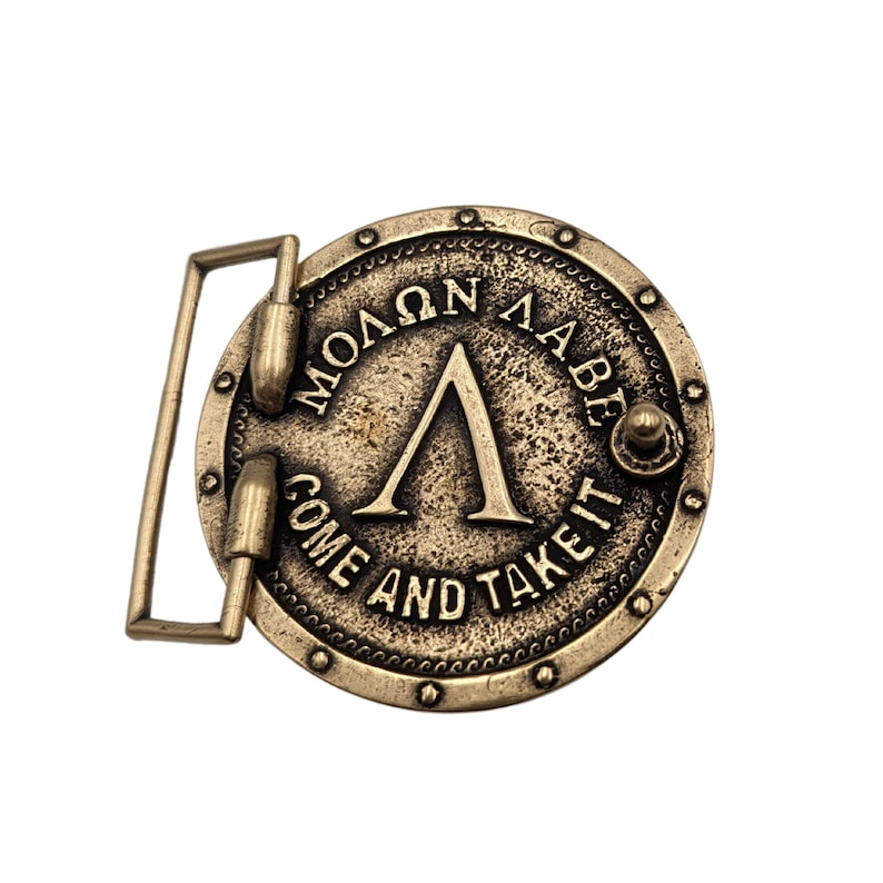 May include: A circular, antique brass-coloured belt buckle featuring a raised Greek Lambda symbol in the centre. The text "&Mu;&Omicron;&Lambda;&Omega;&Nu; &Lambda;&Alpha;&Beta;&Epsilon;" is inscribed above the symbol, with "COME AND TAKE IT" written below. The edge has a riveted, shield-like design.