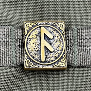 May include: A square, antique gold-colored metal button with a rune symbol in the center. The rune is framed by a circular design and ornate corner patterns. The button is set against a textured, olive-green fabric.