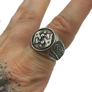 May include: A silver ring with a detailed design of two birds facing each other. The ring is worn on a finger with a tattoo.
