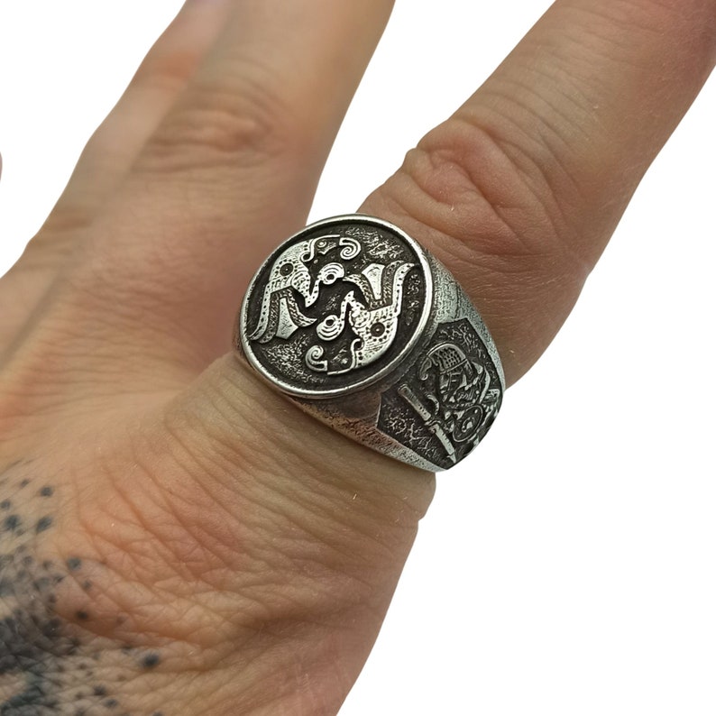 May include: A silver ring with a detailed design of two birds facing each other. The ring is worn on a finger with a tattoo.