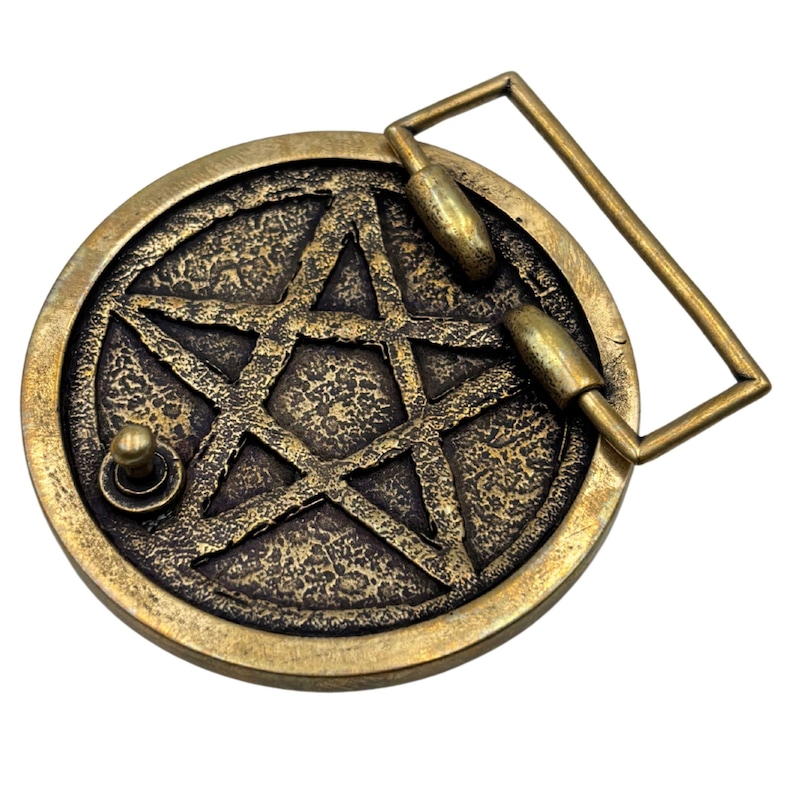 May include: A circular brass-toned belt buckle featuring a raised five-pointed star pentagram design set against a textured, dark background. The buckle includes a rectangular frame attachment and a small metal stud on the front surface.