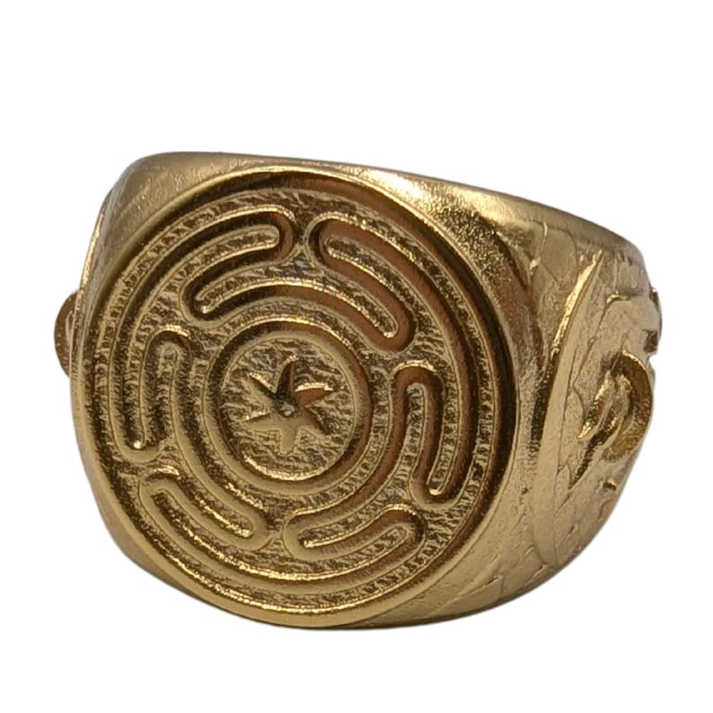 May include: A gold-toned ring featuring a circular face with an embossed labyrinth pattern and a central six-pointed star motif. The band displays textured detailing along the sides, with a polished finish across the entire surface of the ring.