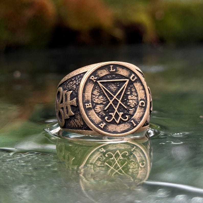 May include: A bronze-toned metal ring featuring a circular face with the engraved text LUCIFER and a central sigil. The band has a textured, dark finish with a cross-like symbol on the side, showcasing a weathered, antique appearance.