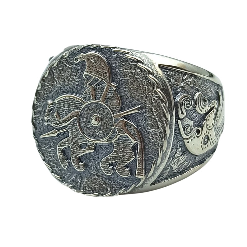May include: A silver ring with a detailed design of a warrior on horseback, holding a spear and shield. The ring has a textured surface and a circular design.
