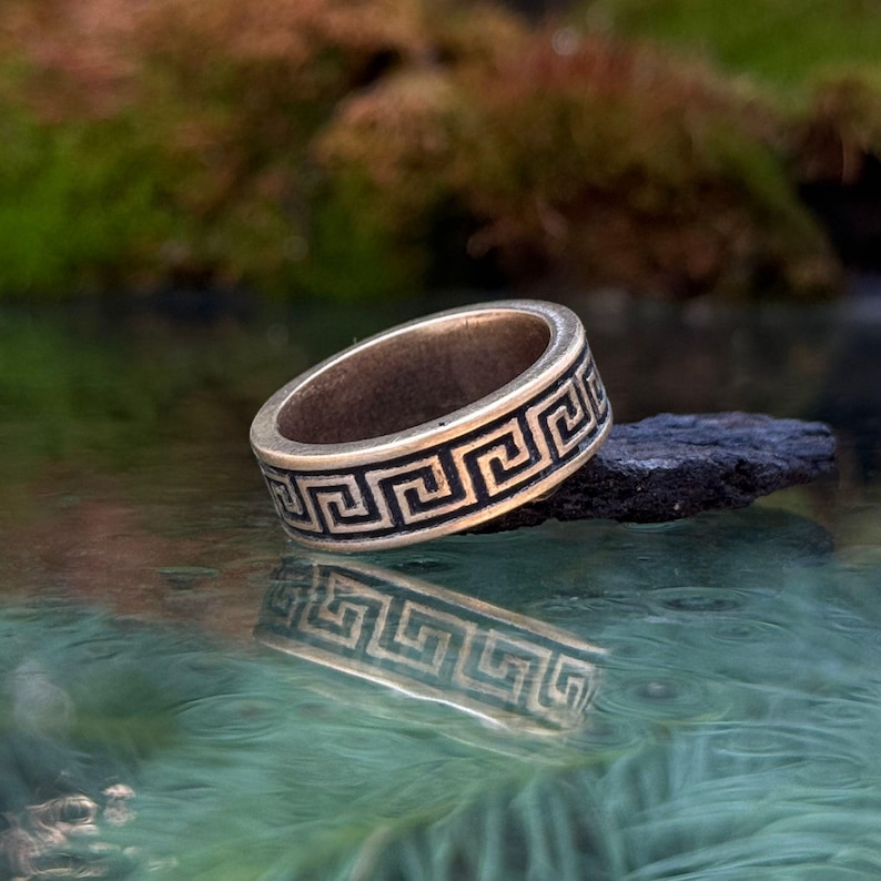 May include: A metallic band ring featuring a continuous black Greek key pattern engraved around the exterior surface. The ring has a weathered, antique finish and is positioned on a dark, textured object above a reflective, rippled surface.