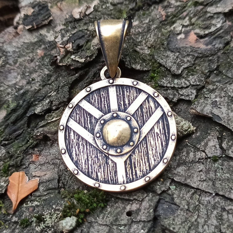 May include: A round, bronze pendant shaped like a shield with a raised, circular design in the centre. The shield has a textured surface and a loop at the top for hanging.