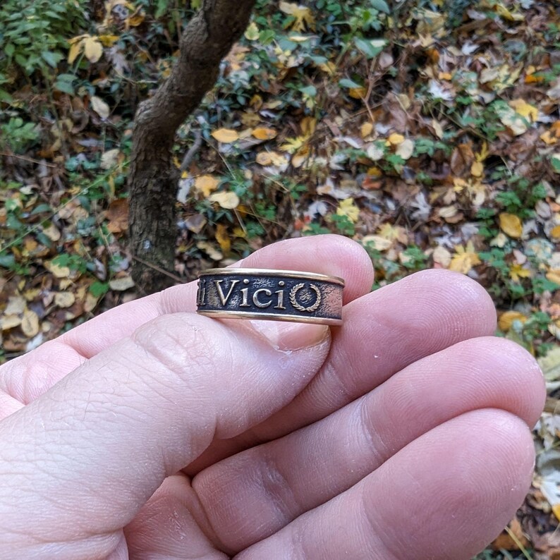 May include: A metallic band ring with a dark, textured finish featuring the embossed text Vici and a small laurel wreath symbol. The ring has smooth, raised edges in a lighter metallic tone, creating a contrast against the recessed, darkened centre.