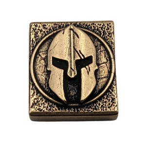 May include: A square, bronze-colored metal tile with a raised, circular design. The design features a Spartan helmet in a black and gold color scheme.