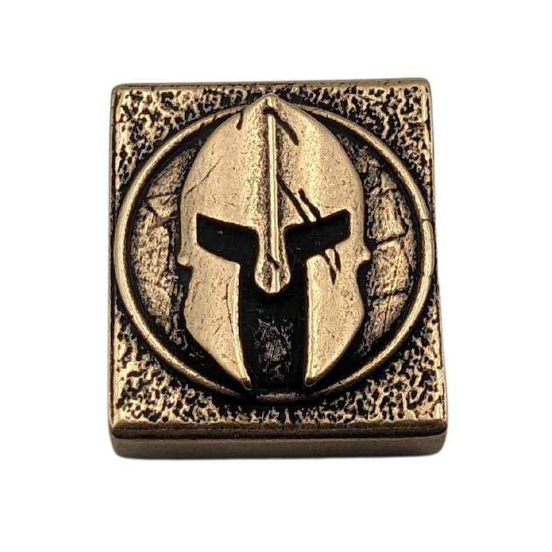 May include: A square, bronze-colored metal tile with a raised, circular design. The design features a Spartan helmet in a black and gold color scheme.