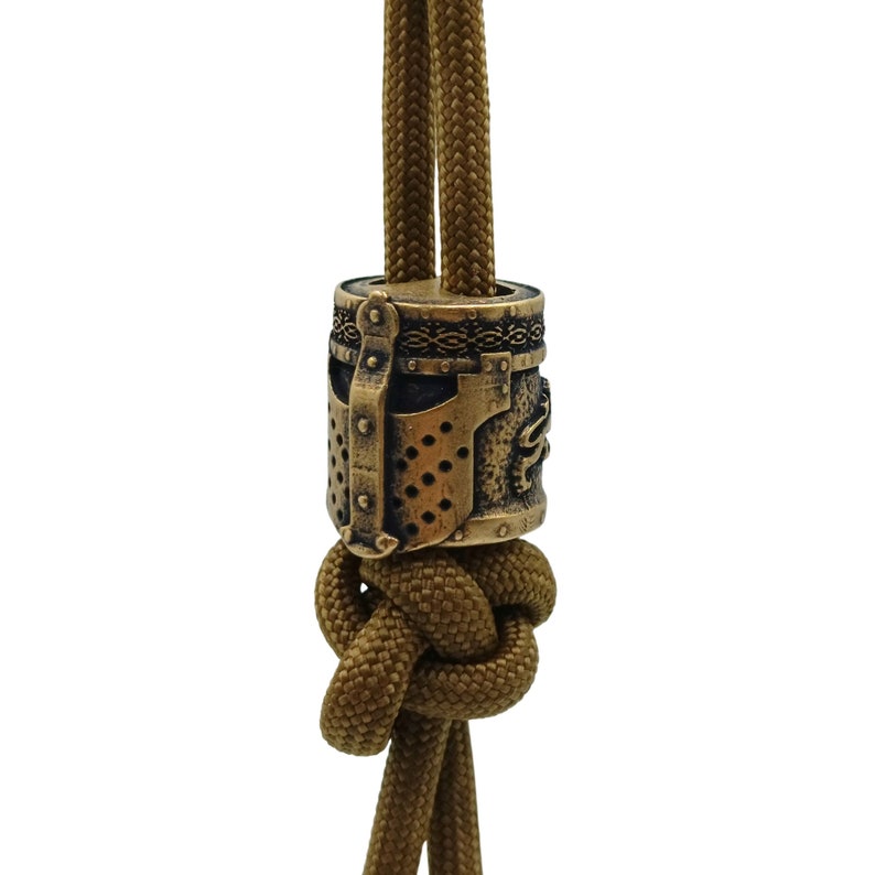 May include: A gold-toned metal bead with a detailed design of a knight's helmet. The bead is strung on a brown paracord.