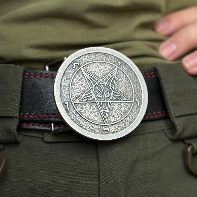 May include: A circular silver-toned metal belt buckle featuring an embossed pentagram design with a central goat head motif and Hebrew characters around the perimeter, attached to a black leather belt with visible red stitching details.