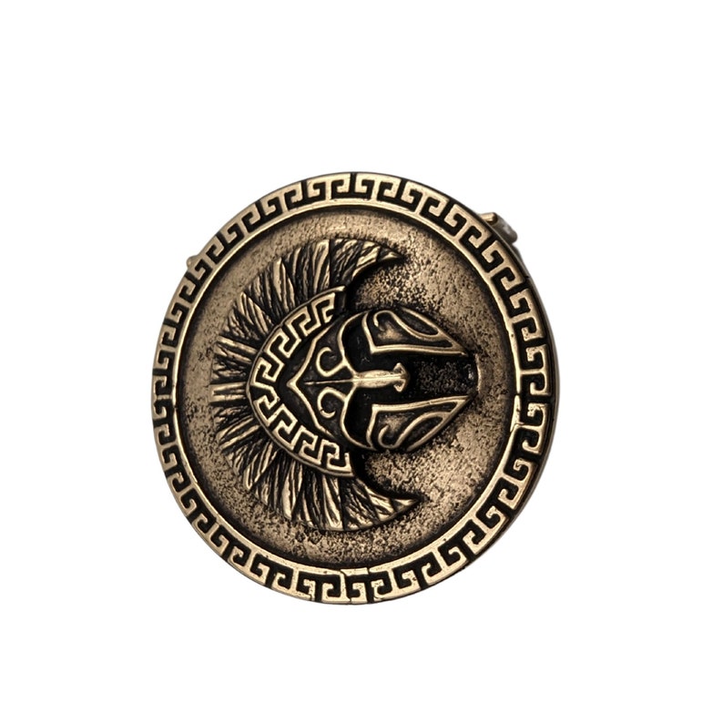 May include: A circular bronze-toned belt buckle featuring a central embossed Spartan helmet design. The outer edge is decorated with a repeating Greek key pattern. The metal surface has a weathered, antiqued finish with dark recessed details.