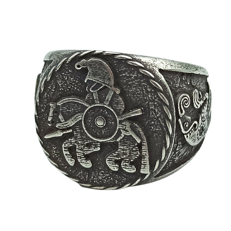 May include: A silver ring with a detailed design of a warrior on horseback. The ring has a rope-like border and a stylized wave design on the side.